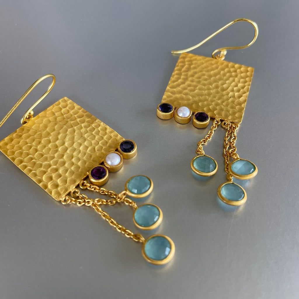 A revamped version of 'Amaya' earrings that was originally launched in 2018 is here!  The design was loved and appreciated by clients a lot. It had a matching ring, bracelets, and a necklace. Since the design did so well it only made sense to add a little twist to it and bring it back. Hence, Athena was created.  3 inches in length and 1 inch in width, the hand-hammered square plate graciously holds 2 amethyst gemstones and a pearl along with 3 blue chalcedonies hung durably via a sleek chain.